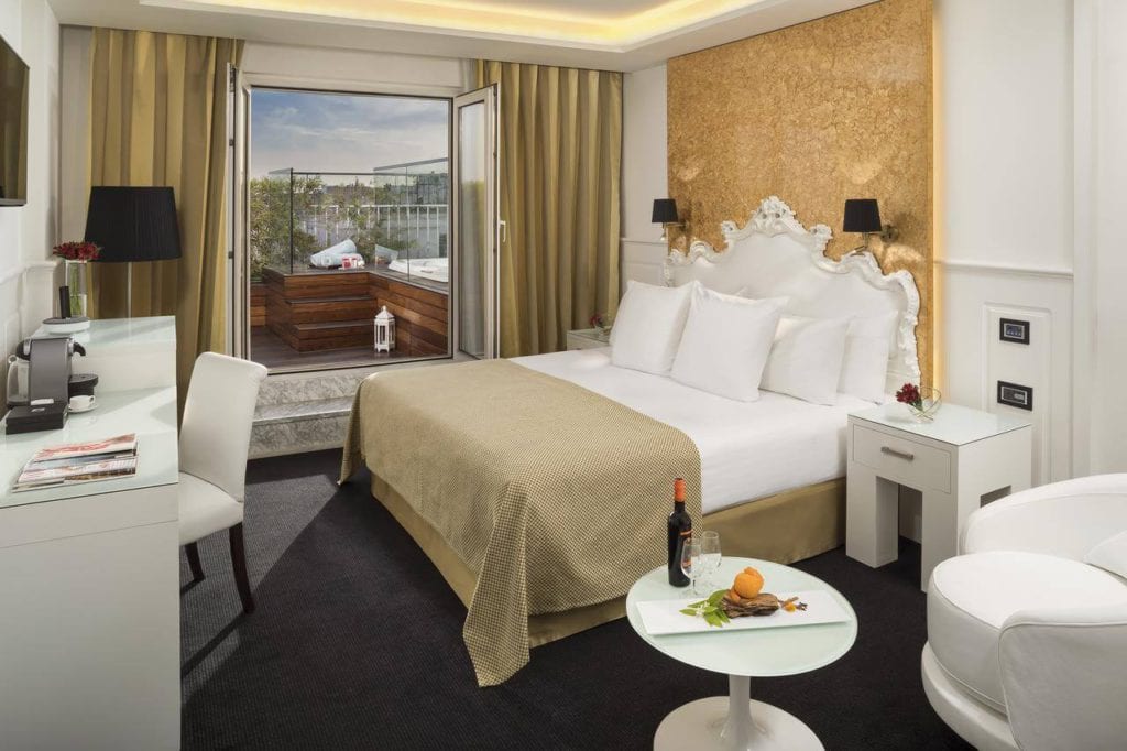 Disfrute de la elegancia del Hotel Gran Meliá Colón Sevilla con una lujosa cama, vista al balcón y una acogedora sala de estar con una mesa decorada con vino y naranjas frescas para mejorar su estadía. - ForTwo - Planes para parejas