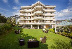 Descubra una escapada romántica en la Escapada romántica al Gran Hotel Victoria con botella de cava (Santander), que cuenta con una gran fachada blanca, encantadores balcones, asientos al aire libre y un exuberante césped verde bajo un cielo despejado. - ForTwo - Planes para parejas