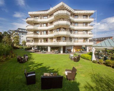 Descubra una escapada romántica en la Escapada romántica al Gran Hotel Victoria con botella de cava (Santander), que cuenta con una gran fachada blanca, encantadores balcones, asientos al aire libre y un exuberante césped verde bajo un cielo despejado. - ForTwo - Planes para parejas