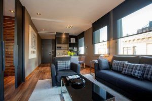 Una moderna sala de estar con sofás oscuros, una mesa de centro de cristal, zona de comedor y grandes ventanales que ofrecen una vista de la ciudad que recuerda a la Grand Suite: la joya de la corona del Grand Hotel Central de Barcelona. - ForTwo - Planes para parejas