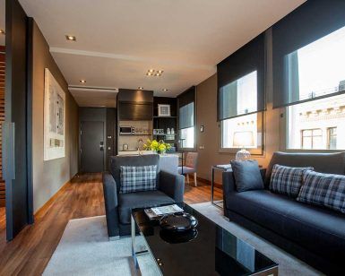 Una moderna sala de estar con sofás oscuros, una mesa de centro de cristal, zona de comedor y grandes ventanales que ofrecen una vista de la ciudad que recuerda a la Grand Suite: la joya de la corona del Grand Hotel Central de Barcelona. - ForTwo - Planes para parejas