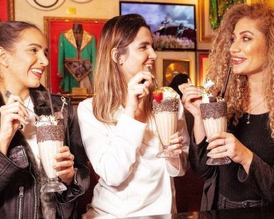 Tres mujeres saborean batidos en el Hard Rock Café Barcelona, sonriendo y charlando en medio de un ambiente bellamente decorado. - ForTwo - Planes para parejas