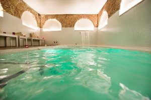 Descubra el Spa Privado con Comida o Cena en el Hospes Puerta de Alcalá, Madrid, con una piscina interior de agua turquesa y arcos de ladrillo. Lujosas tumbonas y velas titilantes crean un ambiente tranquilo en este exclusivo refugio. - ForTwo - Planes para parejas