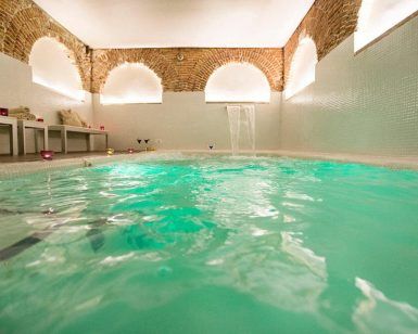 Descubra el Spa Privado con Comida o Cena en el Hospes Puerta de Alcalá, Madrid, con una piscina interior de agua turquesa y arcos de ladrillo. Lujosas tumbonas y velas titilantes crean un ambiente tranquilo en este exclusivo refugio. - ForTwo - Planes para parejas