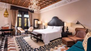 El Hotel Alfonso XIII Sevilla cuenta con un elegante dormitorio con lámpara de araña, cama de gran tamaño y suelo estampado, complementado con muebles clásicos y tonos neutros. - ForTwo - Planes para parejas