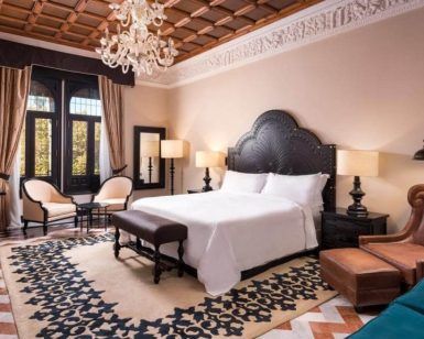 El Hotel Alfonso XIII Sevilla cuenta con un elegante dormitorio con lámpara de araña, cama de gran tamaño y suelo estampado, complementado con muebles clásicos y tonos neutros. - ForTwo - Planes para parejas