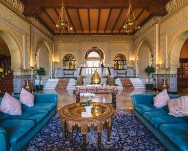 El opulento interior con una decoración ornamentada, sofás azules y una mesa redonda central se asemeja al elegante Hotel Alhambra Palace Granada, con entradas arqueadas que reflejan la grandeza de Granada. - ForTwo - Planes para parejas
