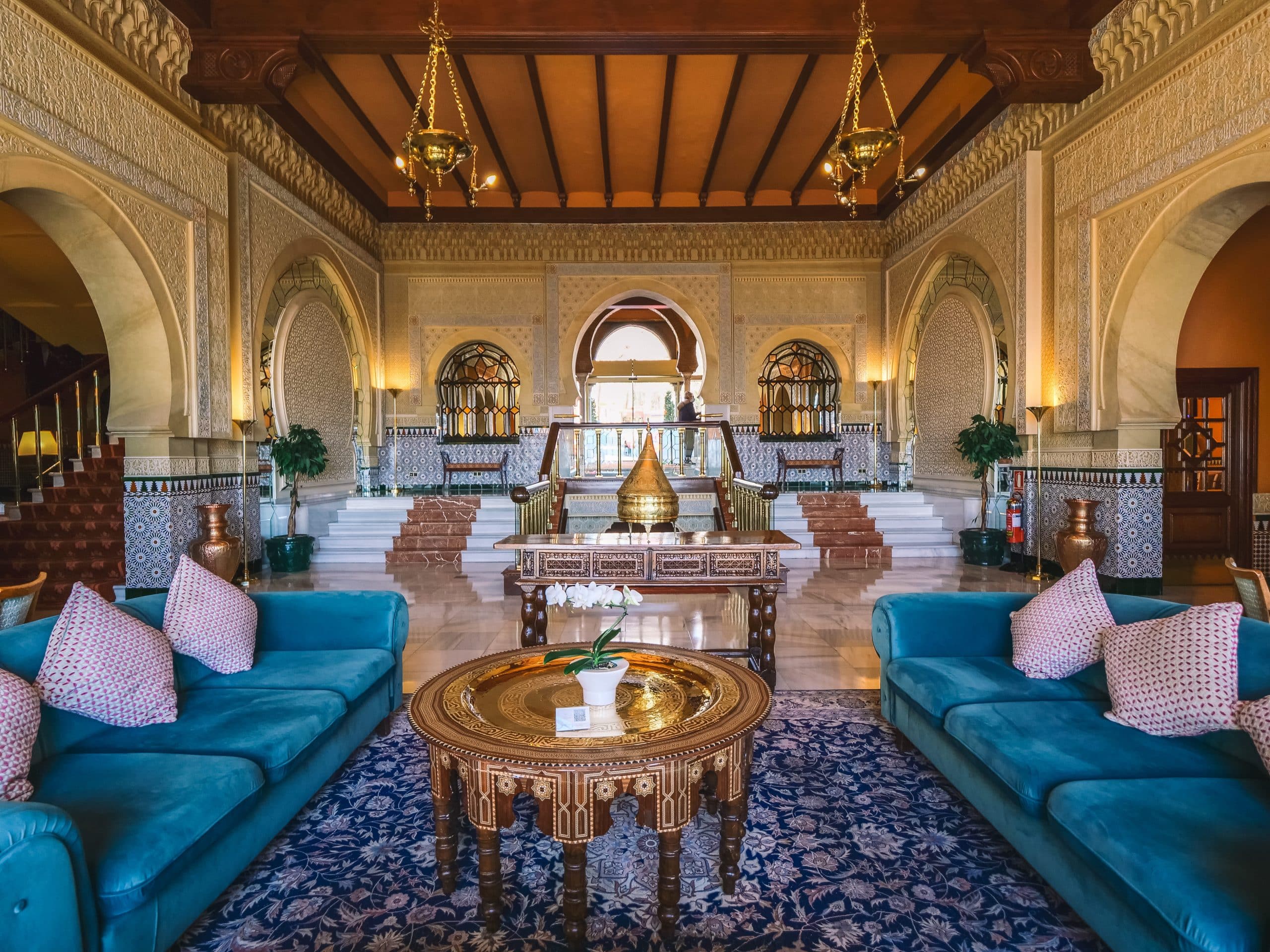 El opulento interior con una decoración ornamentada, sofás azules y una mesa redonda central se asemeja al elegante Hotel Alhambra Palace Granada, con entradas arqueadas que reflejan la grandeza de Granada. - ForTwo - Planes para parejas
