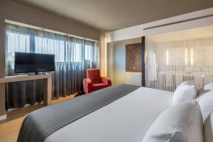 Disfrute de la comodidad del Hotel Barceló Málaga con una lujosa cama king, un cómodo sillón rojo y un elegante televisor de pantalla plana. El elegante baño con paredes de cristal esmerilado realzará su estancia en la vibrante Málaga. - ForTwo - Planes para parejas