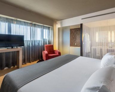 Disfrute de la comodidad del Hotel Barceló Málaga con una lujosa cama king, un cómodo sillón rojo y un elegante televisor de pantalla plana. El elegante baño con paredes de cristal esmerilado realzará su estancia en la vibrante Málaga. - ForTwo - Planes para parejas
