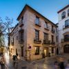 En una plaza adoquinada, ciclistas y peatones se mueven entre históricos edificios de ladrillo. El encantador Hotel Boutique Gar Anat Granada añade un toque del rico patrimonio de la ciudad a la pintoresca escena callejera nocturna. - ForTwo - Planes para parejas