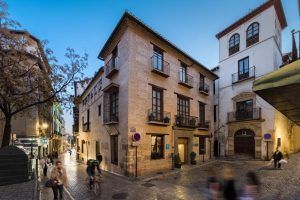En una plaza adoquinada, ciclistas y peatones se mueven entre históricos edificios de ladrillo. El encantador Hotel Boutique Gar Anat Granada añade un toque del rico patrimonio de la ciudad a la pintoresca escena callejera nocturna. - ForTwo - Planes para parejas
