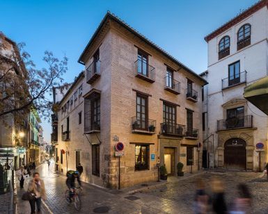 En una plaza adoquinada, ciclistas y peatones se mueven entre históricos edificios de ladrillo. El encantador Hotel Boutique Gar Anat Granada añade un toque del rico patrimonio de la ciudad a la pintoresca escena callejera nocturna. - ForTwo - Planes para parejas