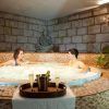 Dos personas se relajan en el jacuzzi del spa durante el "Fin de semana romántico en Hotel Spa Cádiz Playa", con champán en una bandeja a su lado y una pared de piedra realzando la atmósfera serena. - ForTwo - Planes para parejas