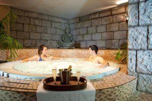 Dos personas se relajan en el jacuzzi del spa durante el "Fin de semana romántico en Hotel Spa Cádiz Playa", con champán en una bandeja a su lado y una pared de piedra realzando la atmósfera serena. - ForTwo - Planes para parejas