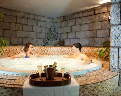 Dos personas se relajan en el jacuzzi del spa durante el "Fin de semana romántico en Hotel Spa Cádiz Playa", con champán en una bandeja a su lado y una pared de piedra realzando la atmósfera serena. - ForTwo - Planes para parejas