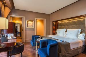 Elegante habitación en el Hotel Casa Fuster GL Monumento en Barcelona con una cama grande, lujosas sillas azules, escritorio de madera, iluminación cálida; las puertas evocan el encanto de la arquitectura del Monumento. - ForTwo - Planes para parejas