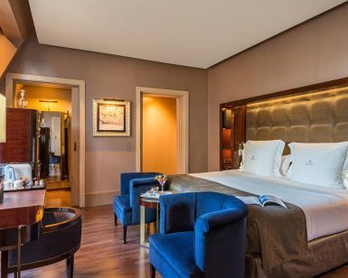 Elegante habitación en el Hotel Casa Fuster GL Monumento en Barcelona con una cama grande, lujosas sillas azules, escritorio de madera, iluminación cálida; las puertas evocan el encanto de la arquitectura del Monumento. - ForTwo - Planes para parejas