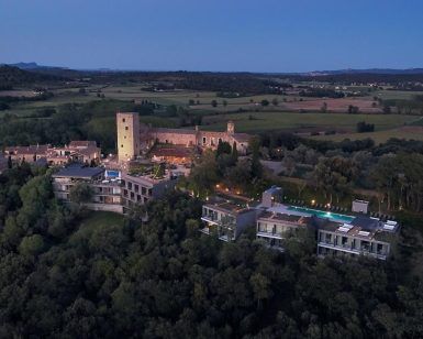 Vista aérea del Hotel Castell d'Emporda (Girona) iluminado y estructuras cercanas enmarcadas en el paisaje rural al anochecer. - ForTwo - Planes para parejas