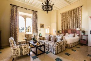 Una elegante habitación en el Hotel Castillo de Santa Catalina Málaga cuenta con una cama doble, muebles con motivos florales, una lámpara de araña y ventanas altas con cortinas largas. - ForTwo - Planes para parejas