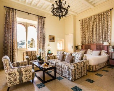 Una elegante habitación en el Hotel Castillo de Santa Catalina Málaga cuenta con una cama doble, muebles con motivos florales, una lámpara de araña y ventanas altas con cortinas largas. - ForTwo - Planes para parejas