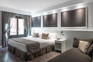 Descubra el encanto de Barcelona en las modernas habitaciones del Hotel Catalonia Magdalenes, que cuentan con una cama grande, paneles de pared decorativos, un acogedor sofá, ventanas con cortinas y elegantes mesitas de noche. - ForTwo - Planes para parejas
