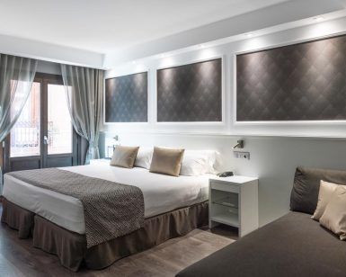Descubra el encanto de Barcelona en las modernas habitaciones del Hotel Catalonia Magdalenes, que cuentan con una cama grande, paneles de pared decorativos, un acogedor sofá, ventanas con cortinas y elegantes mesitas de noche. - ForTwo - Planes para parejas