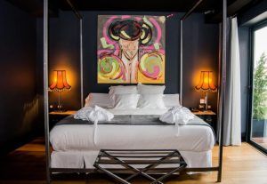 La habitación del vestíbulo del hotel Sevilla presenta una decoración moderna con una cama con dosel, arte abstracto vibrante y dos lámparas naranjas que iluminan las paredes oscuras, creando un ambiente acogedor. - ForTwo - Planes para parejas