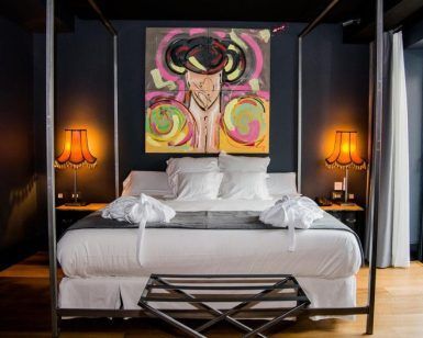 La habitación del vestíbulo del hotel Sevilla presenta una decoración moderna con una cama con dosel, arte abstracto vibrante y dos lámparas naranjas que iluminan las paredes oscuras, creando un ambiente acogedor. - ForTwo - Planes para parejas