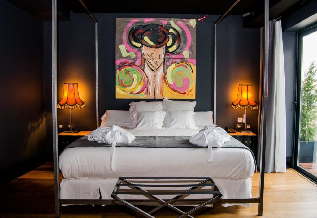La habitación del vestíbulo del hotel Sevilla presenta una decoración moderna con una cama con dosel, arte abstracto vibrante y dos lámparas naranjas que iluminan las paredes oscuras, creando un ambiente acogedor. - ForTwo - Planes para parejas