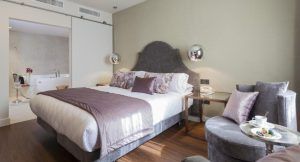 Descubra la elegancia del Hotel Palacete de Álamos Málaga: disfrute de una elegante habitación con una cama tamaño king, detalles en morado, cómodas sillas y un moderno baño abierto con una lujosa bañera para la combinación perfecta de comodidad y sofisticación. - ForTwo - Planes para parejas
