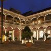 El patio iluminado del Hotel Palacio de Santa Paula Granada brilla cálidamente por la noche, con arcos que crean sombras en los árboles en macetas y un diseño de azulejos central que irradia encanto histórico. - ForTwo - Planes para parejas