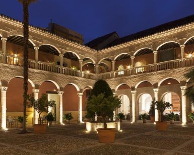 El patio iluminado del Hotel Palacio de Santa Paula Granada brilla cálidamente por la noche, con arcos que crean sombras en los árboles en macetas y un diseño de azulejos central que irradia encanto histórico. - ForTwo - Planes para parejas