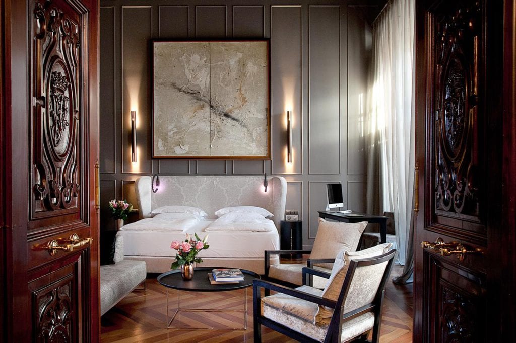 El elegante dormitorio del Hotel Palacio de Villapanés Sevilla incluye dos camas individuales, grandes obras de arte abstracto, puertas de madera ornamentadas y sillas, realzadas por una iluminación suave y flores que le añaden encanto. - ForTwo - Planes para parejas
