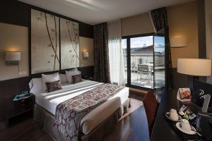 Moderna habitación en el Hotel Paseo Del Arte (Madrid) con una amplia cama y escritorio, además de una puerta de balcón abierta que revela una terraza con impresionantes vistas de la ciudad. - ForTwo - Planes para parejas