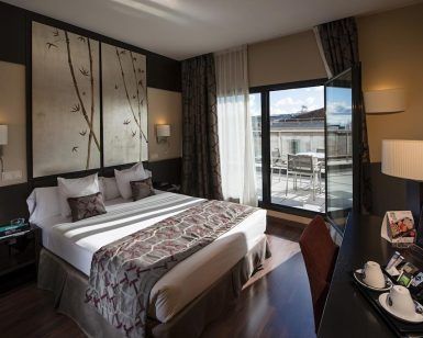 Moderna habitación en el Hotel Paseo Del Arte (Madrid) con una amplia cama y escritorio, además de una puerta de balcón abierta que revela una terraza con impresionantes vistas de la ciudad. - ForTwo - Planes para parejas