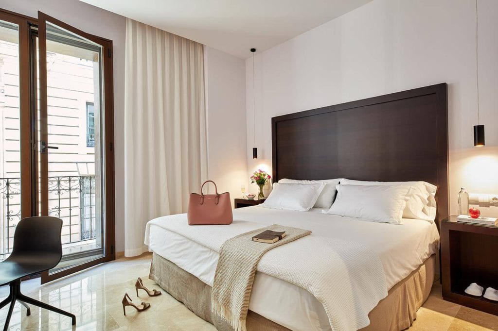Disfrute de una moderna habitación en el Hotel Posada del Lucero Sevilla, con una amplia cama con mesitas de noche, una silla y un acogedor bolso sobre la cama. Las puertas del balcón le permiten disfrutar del animado ambiente sevillano desde su habitación. - ForTwo - Planes para parejas