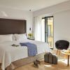 En la radiante habitación del Hotel Rey Alfonso X Sevilla, una espaciosa cama promete relajación, con una mesa que ofrece comodidades para el desayuno y una silla para relajarse. Una maleta junto a la ventana completa el conjunto, encapsulando el encanto de Sevilla. - ForTwo - Planes para parejas