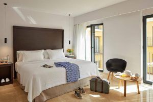 En la radiante habitación del Hotel Rey Alfonso X Sevilla, una espaciosa cama promete relajación, con una mesa que ofrece comodidades para el desayuno y una silla para relajarse. Una maleta junto a la ventana completa el conjunto, encapsulando el encanto de Sevilla. - ForTwo - Planes para parejas