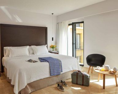 En la radiante habitación del Hotel Rey Alfonso X Sevilla, una espaciosa cama promete relajación, con una mesa que ofrece comodidades para el desayuno y una silla para relajarse. Una maleta junto a la ventana completa el conjunto, encapsulando el encanto de Sevilla. - ForTwo - Planes para parejas