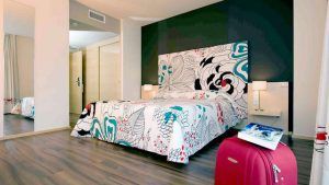 Una moderna habitación de hotel con una cama con estampado floral, una pared con acento oscuro, un piso de madera y una maleta roja prepara el escenario para una 'Escapada 50 Sombras de gris con Spa, lovebox, Jacuzzi privado (Murcia)', que ofrece una estadía intrigante y lujosa. - ForTwo - Planes para parejas