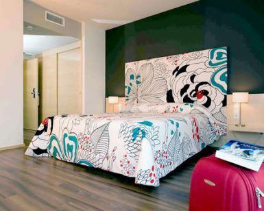 Una moderna habitación de hotel con una cama con estampado floral, una pared con acento oscuro, un piso de madera y una maleta roja prepara el escenario para una 'Escapada 50 Sombras de gris con Spa, lovebox, Jacuzzi privado (Murcia)', que ofrece una estadía intrigante y lujosa. - ForTwo - Planes para parejas