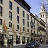 Un edificio europeo con encantadoras tiendas y banderas se alza junto a un coche aparcado. La histórica torre de la iglesia de Granada se alza al fondo, junto al elegante Hotel Vincci Centro Granada, creando un pintoresco paisaje urbano. - ForTwo - Planes para parejas