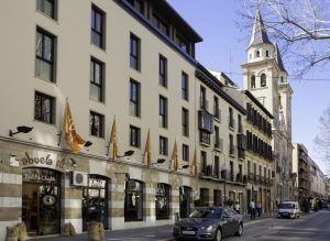 Un edificio europeo con encantadoras tiendas y banderas se alza junto a un coche aparcado. La histórica torre de la iglesia de Granada se alza al fondo, junto al elegante Hotel Vincci Centro Granada, creando un pintoresco paisaje urbano. - ForTwo - Planes para parejas
