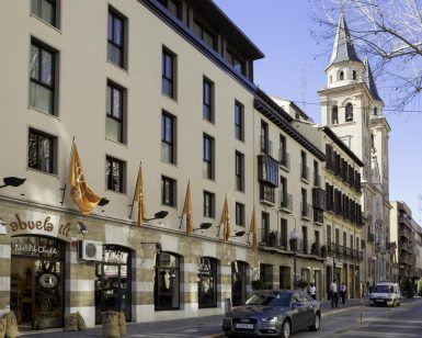 Un edificio europeo con encantadoras tiendas y banderas se alza junto a un coche aparcado. La histórica torre de la iglesia de Granada se alza al fondo, junto al elegante Hotel Vincci Centro Granada, creando un pintoresco paisaje urbano. - ForTwo - Planes para parejas