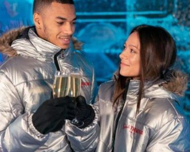 Dentro de ICEBARCELONA, una pareja con chaquetas plateadas choca sus copas, sus sonrisas iluminan el fondo helado de la experiencia Pack Romántico en ICEBARCELONA. - ForTwo - Planes para parejas