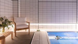 La acogedora piscina cubierta, con elegantes paredes de azulejos blancos, refleja el ambiente del Gran Hotel Inglés. Una silla de madera con cojines y un banco adornado con flores crean un remanso de paz ideal para relajarse después de disfrutar de un jacuzzi y un masaje privado en este distinguido hotel madrileño. - ForTwo - Planes para parejas