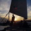 Silueta de personas en el Crucero en catamarán con Puesta de Sol en Barcelona, uno tocando el saxofón, con vela y agua tranquila al fondo. - ForTwo - Planes para parejas