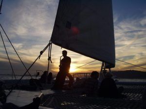 Silueta de personas en el Crucero en catamarán con Puesta de Sol en Barcelona, uno tocando el saxofón, con vela y agua tranquila al fondo. - ForTwo - Planes para parejas