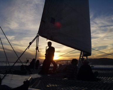 Silueta de personas en el Crucero en catamarán con Puesta de Sol en Barcelona, uno tocando el saxofón, con vela y agua tranquila al fondo. - ForTwo - Planes para parejas